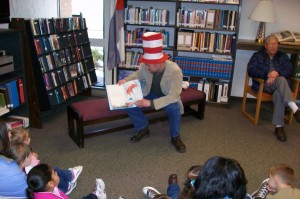 man with Cat In the Hat hat on reads a Dr. Seuss book to children